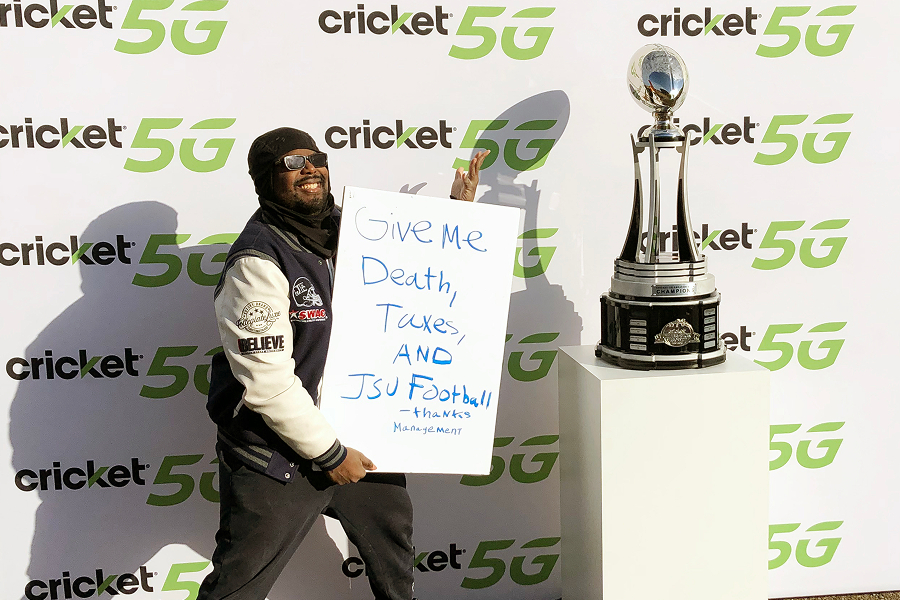 JSU fan holding a humorous sign reading 'Give Me Death, Taxes, AND JSU Football' next to the Cricket Celebration Bowl Champions trophy at a Cricket 5G activation