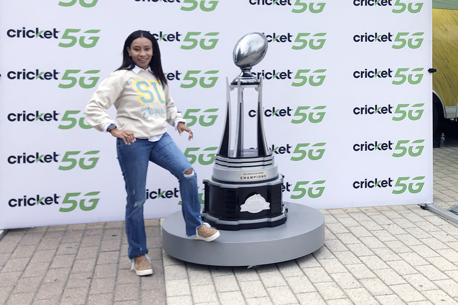Southern University fan in SU Jaguars gear posing confidently next to the Cricket Celebration Bowl Champions trophy at a Cricket 5G activation